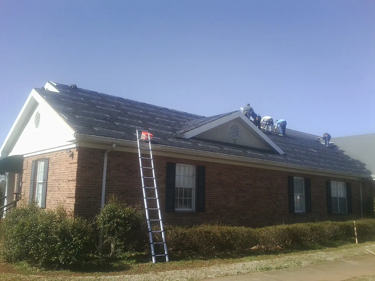 Crew tearing shingles off a brick ranch roof with a ladder set — Emergency Roof Repair work by our crew in Lenoir