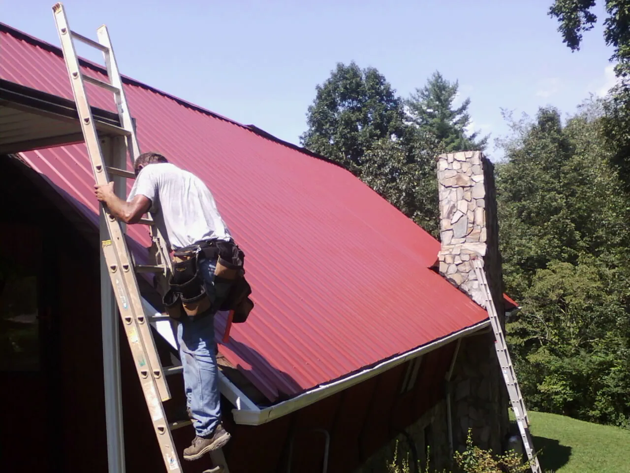 Roofer on a ladder beside a red metal roof and stone chimney — Soffit & Fascia Repair by Roofing Brothers in Lenoir
